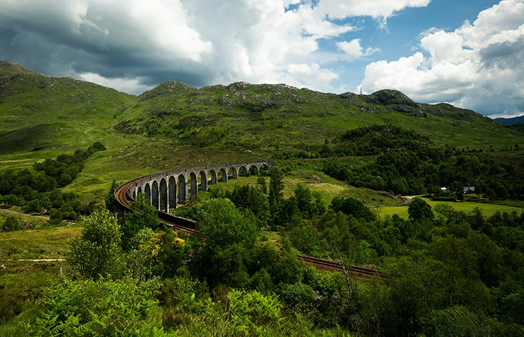 A scenic view of a viaduct with multiple arches, spanning across a lush green valley and surrounded by rolling hills under a partly cloudy sky.