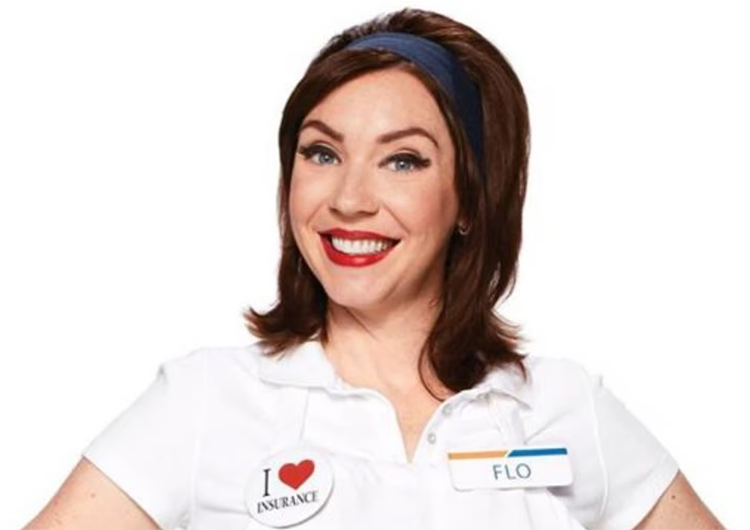 A woman with shoulder-length brown hair, wearing a white shirt and headband, smiles. She has a name tag reading "FLO" and a badge with "I ❤️ INSURANCE" on her chest.