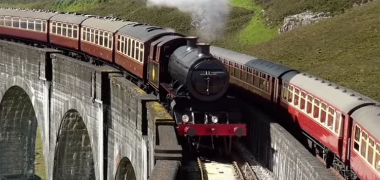 Steam locomotive with a plume of smoke pulling vintage red and cream passenger cars, traveling on a curved stone viaduct surrounded by green hills.