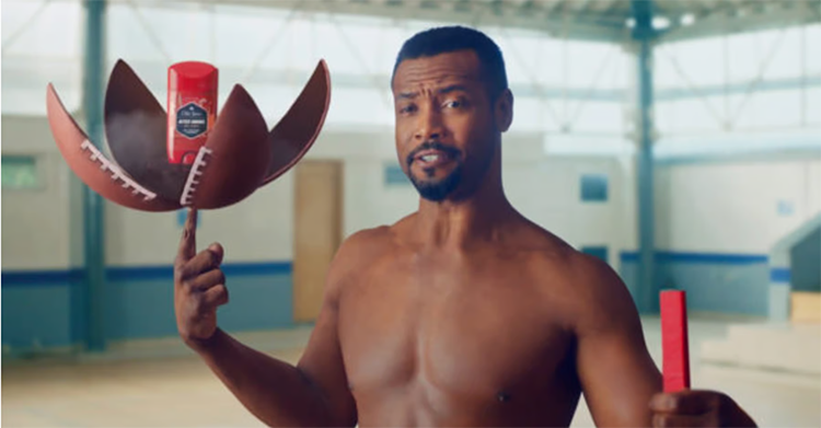 A shirtless man standing indoors holding a split football with a deodorant inside in one hand and a red stick in the other hand.